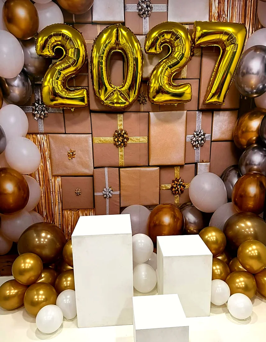 Cenário de Ano-Novo: balões dourados formando 2027, painel de presentes e cubos brancos